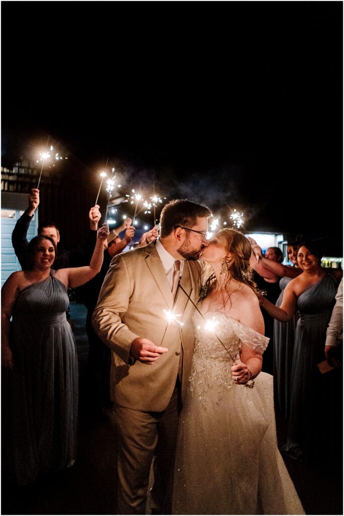 Snohomish wedding photographer gsquared weddings a bride and groom kiss while holding sparklers surrounded by cheering guests also holding sparklers The bride wears an off the shoulder gown and the groom is in a light suit The scene is nighttime creating a festive illuminated atmosphere Serving seattle snohomish and washington state