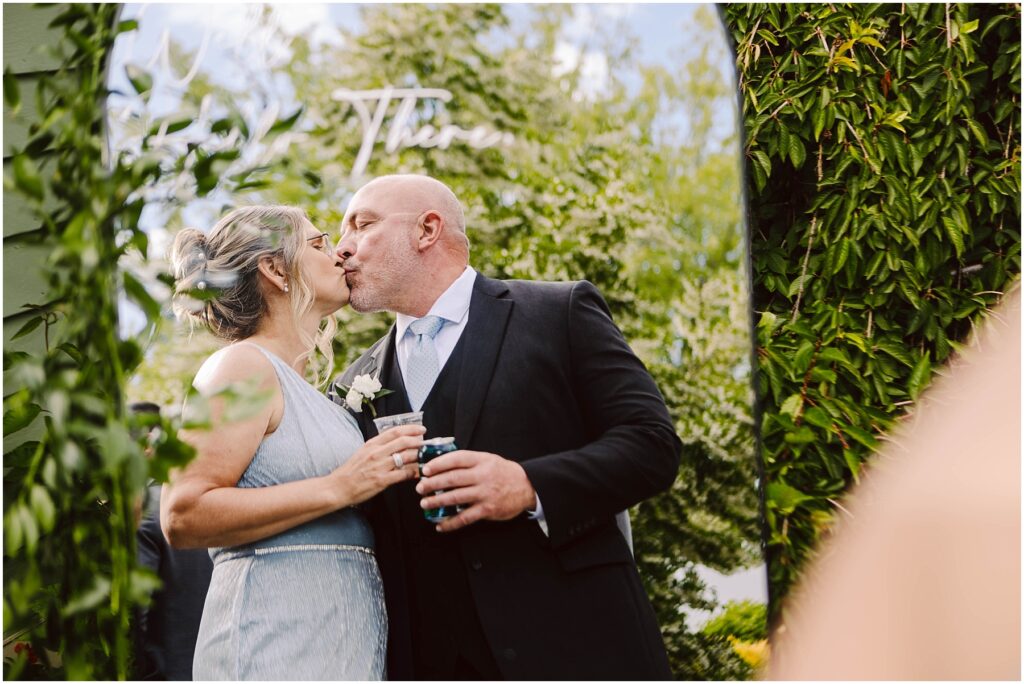 Snohomish Wedding Photographer GSquared Weddings A couple dressed formally shares a kiss in an outdoor setting with lush greenery. The woman holds a drink and wears a light blue dress, while the man, in a suit, holds a beverage can. The background features trees and foliage. Serving Seattle, Snohomish and Washington State