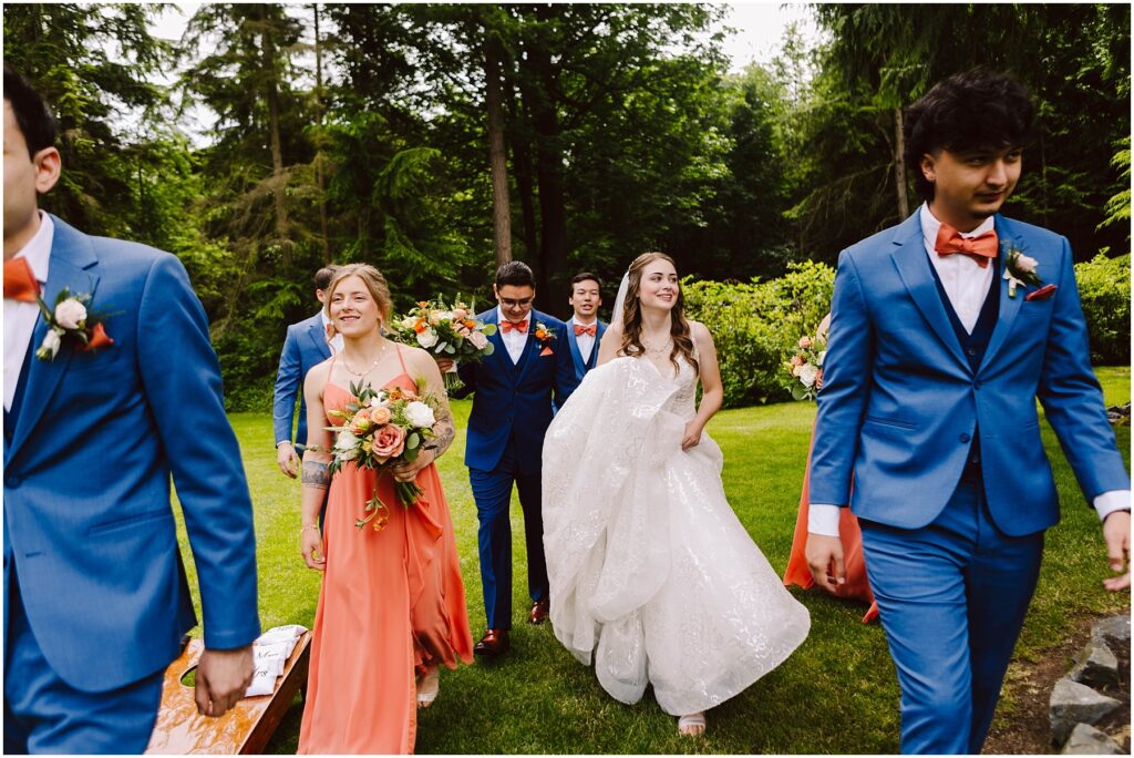 Snohomish Wedding Photographer GSquared Weddings A bride in a white dress walks joyfully with a groom and bridal party through a lush garden. The groomsmen wear blue suits with orange bow ties, and the bridesmaids wear orange dresses, holding bouquets of flowers. Serving Seattle, Snohomish and Washington State