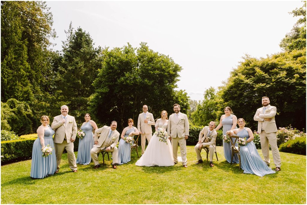 Snohomish Wedding Photographer GSquared Weddings A wedding party poses outdoors on a grassy lawn. The bride and groom stand in the center, surrounded by bridesmaids in light blue dresses and groomsmen in beige suits. Trees and greenery form the backdrop under a sunny sky. Serving Seattle, Snohomish and Washington State