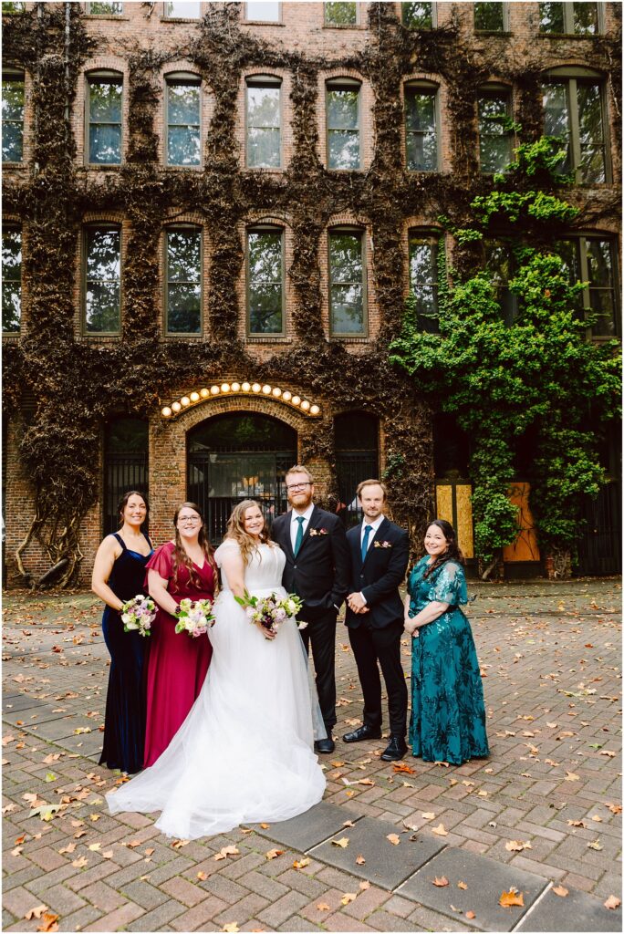 Snohomish Wedding Photographer GSquared Weddings A wedding group poses outdoors in front of a historic brick building with ivy. The bride stands with a groom and four bridesmaids holding bouquets. The scene is set on a cobblestone path with scattered autumn leaves. Serving Seattle, Snohomish and Washington State
