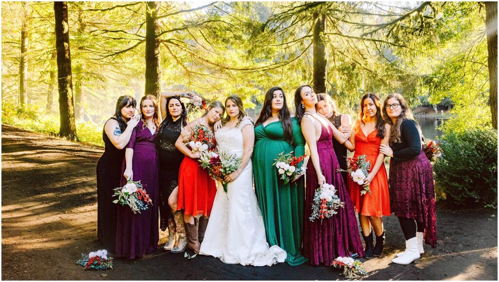 Snohomish Wedding Photographer GSquared Weddings A group of women, including a bride in a white gown, stand in a forest, surrounded by sunlight filtering through the trees. They wear colorful dresses in various shades, holding bouquets. The scene is warm and vibrant with lush greenery. Serving Seattle, Snohomish and Washington State