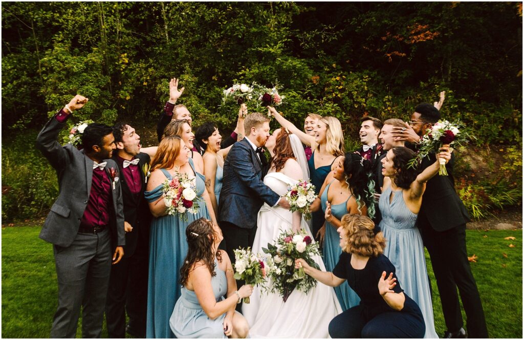 Snohomish Wedding Photographer GSquared Weddings A wedding party poses outdoors with the bride and groom kissing in the center. Bridesmaids in blue dresses and groomsmen in dark suits cheer and hold bouquets against a green, leafy background. Everyone appears joyful and celebratory. Serving Seattle, Snohomish and Washington State