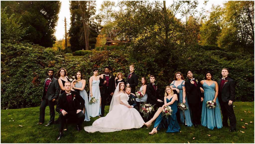 Snohomish Wedding Photographer GSquared Weddings A wedding party poses outdoors on a lawn against a backdrop of trees and greenery. The bride sits in the center in a white gown, surrounded by bridesmaids in blue dresses and groomsmen in dark suits. The atmosphere is elegant and relaxed. Serving Seattle, Snohomish and Washington State