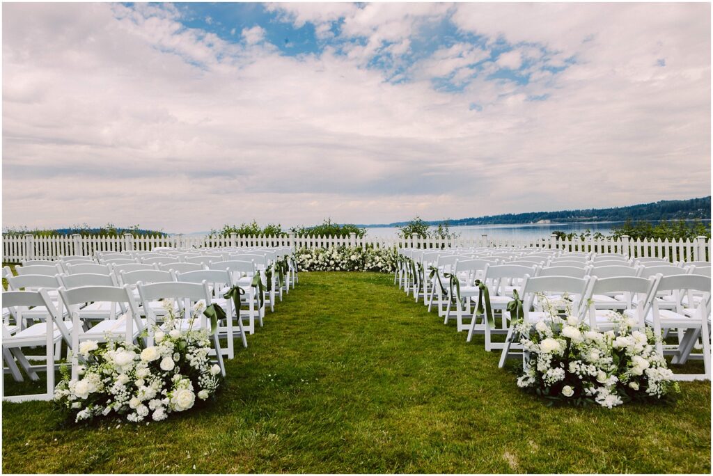 Outdoor wedding ceremony setup with white folding chairs in rows white floral arrangements lining the grass aisle overlooking water and a white picket fence Snohomish wedding photographer gsquared weddings a scenic outdoor wedding setup features two rows of white chairs lined with white floral arrangements on a grassy area A white picket fence overlooks a body of water and a cloudy sky providing a picturesque backdrop Serving seattle snohomish and washington state
