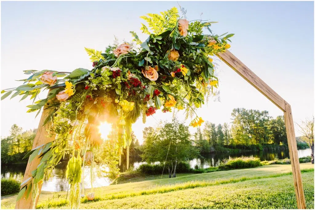 Snohomish Wedding Photographer GSquared Weddings A wooden arch adorned with green leaves pink and yellow flowers with sunlight shining through The arch is set in a grassy area near a calm river and trees are visible in the background under a bright blue sky Serving Seattle Snohomish and Washington State Snohomish Wedding Photographer GSquared Weddings A wooden arch adorned with green leaves, pink and yellow flowers, with sunlight shining through. The arch is set in a grassy area near a calm river, and trees are visible in the background under a bright blue sky. Serving Seattle, Snohomish and Washington State