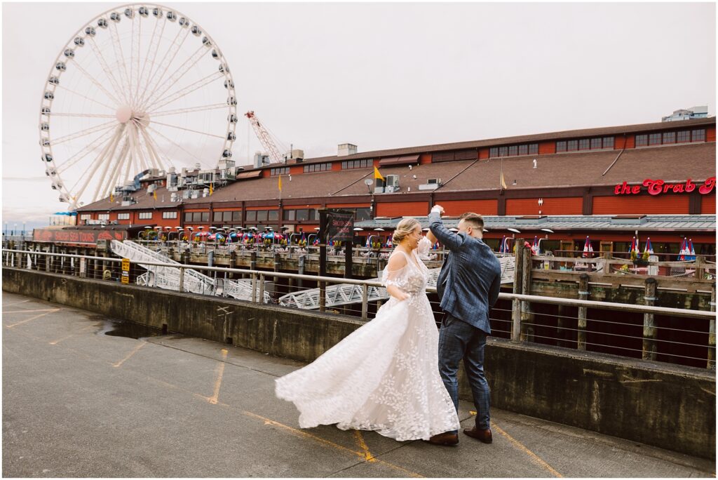 Bride and groom dancing on a waterfront pier with the seattle great wheel in the background Snohomish wedding photographer gsquared weddings a couple dances outside near a waterfront The woman wears a white wedding dress and the man is in a blue suit In the background theres a large ferris wheel and a building with a sign that reads the crab pot Serving seattle snohomish and washington state