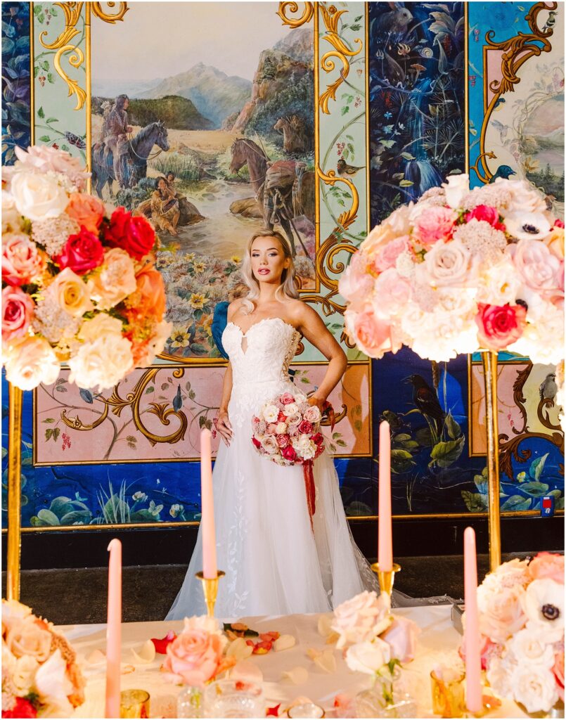 Snohomish wedding photographer gsquared weddings a bride in a white gown holding a bouquet stands in front of a detailed mural depicting scenes and landscapes The foreground features flower arrangements with pink and red roses and tall pink candles Serving seattle snohomish and washington state Snohomish wedding photographer gsquared weddings a bride in a white gown holding a bouquet stands in front of a detailed mural depicting scenes and landscapes The foreground features flower arrangements with pink and red roses and tall pink candles Serving seattle snohomish and washington state
