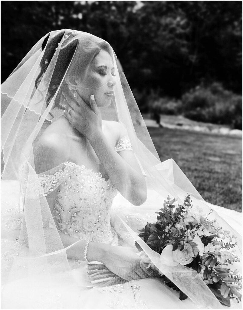 Snohomish wedding photographer gsquared weddings bride in an intricately beaded gown sits outdoors partially covered by a sheer veil She holds a bouquet and rests one hand on her neck looking serene The background features a blurred natural setting Black and white image Serving seattle snohomish and washington state Snohomish wedding photographer gsquared weddings bride in an intricately beaded gown sits outdoors partially covered by a sheer veil She holds a bouquet and rests one hand on her neck looking serene The background features a blurred natural setting Black and white image Serving seattle snohomish and washington state