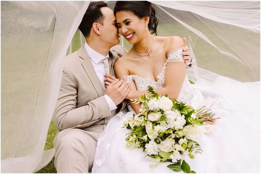 Snohomish wedding photographer gsquared weddings bride and groom sitting under a veil smiling The bride is in an off-shoulder white dress holding a bouquet of white flowers The groom is in a beige suit Theyre on grass sharing an intimate joyful moment Serving seattle snohomish and washington state Snohomish wedding photographer gsquared weddings bride and groom sitting under a veil smiling The bride is in an off shoulder white dress holding a bouquet of white flowers The groom is in a beige suit Theyre on grass sharing an intimate joyful moment Serving seattle snohomish and washington state