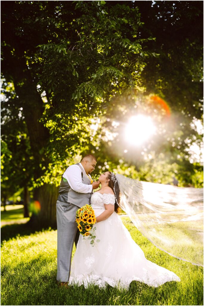 Groom touches the brides chin under a tree as sunlight flares behind them her veil blowing and a sunflower bouquet in hand Snohomish wedding photographer gsquared weddings a bride and groom stand under a large tree with sunlight streaming through the leaves The bride holds a bouquet of sunflowers and her veil flows in the breeze Both are gazing at each other smiling warmly Serving seattle snohomish and washington state