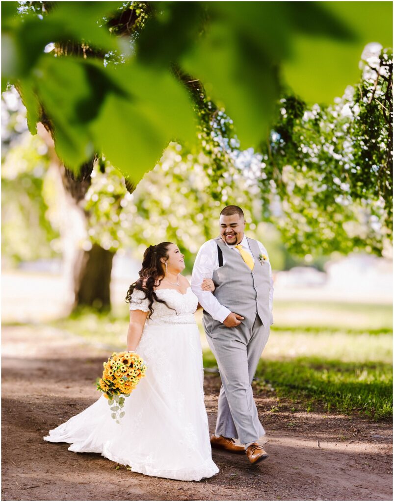 Snohomish wedding photographer gsquared weddings a bride in a white dress holds a sunflower bouquet walking hand in hand with a groom in a gray suit and suspenders They are smiling at each other under lush green trees sharing a joyful moment on a sunlit path Serving seattle snohomish and washington state Snohomish wedding photographer gsquared weddings a bride in a white dress holds a sunflower bouquet walking hand in hand with a groom in a gray suit and suspenders They are smiling at each other under lush green trees sharing a joyful moment on a sunlit path Serving seattle snohomish and washington state