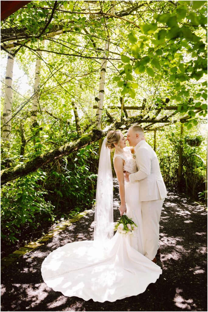 Snohomish wedding photographer gsquared weddings a couple in wedding attire shares a kiss under sun-dappled trees The bride holds a bouquet and wears a long veil while the groom is in a light suit Sunlight filters through the lush green leaves creating a serene and romantic scene Serving seattle snohomish and washington state Snohomish wedding photographer gsquared weddings a couple in wedding attire shares a kiss under sun dappled trees The bride holds a bouquet and wears a long veil while the groom is in a light suit Sunlight filters through the lush green leaves creating a serene and romantic scene Serving seattle snohomish and washington state