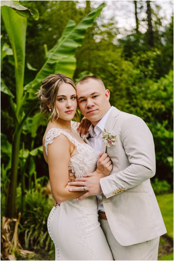 Snohomish wedding photographer gsquared weddings a bride in a white lace dress and a groom in a light gray suit pose together in a lush garden setting They are embracing looking at the camera with large green leaves visible in the background Serving seattle snohomish and washington state Snohomish wedding photographer gsquared weddings a bride in a white lace dress and a groom in a light gray suit pose together in a lush garden setting They are embracing looking at the camera with large green leaves visible in the background Serving seattle snohomish and washington state