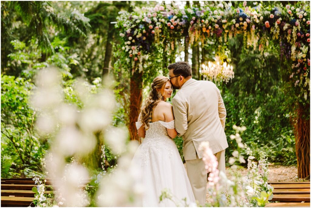 Snohomish wedding photographer gsquared weddings a bride in a white dress and a groom in a beige suit stand embracing under a floral arch in a lush green outdoor setting The groom kisses the brides cheek as they face away from the camera surrounded by vibrant plants and flowers Serving seattle snohomish and washington state Snohomish wedding photographer gsquared weddings a bride in a white dress and a groom in a beige suit stand embracing under a floral arch in a lush green outdoor setting The groom kisses the brides cheek as they face away from the camera surrounded by vibrant plants and flowers Serving seattle snohomish and washington state