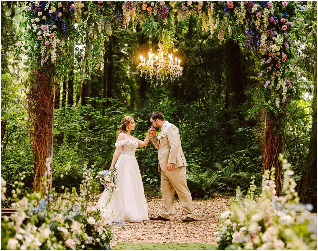 Snohomish wedding photographer gsquared weddings a bride in a white dress and groom in a beige suit stand under a floral arch in a forest The groom kisses the brides hand A chandelier hangs above them and colorful flowers surround the scene Serving seattle snohomish and washington state Snohomish wedding photographer gsquared weddings a bride in a white dress and groom in a beige suit stand under a floral arch in a forest The groom kisses the brides hand A chandelier hangs above them and colorful flowers surround the scene Serving seattle snohomish and washington state