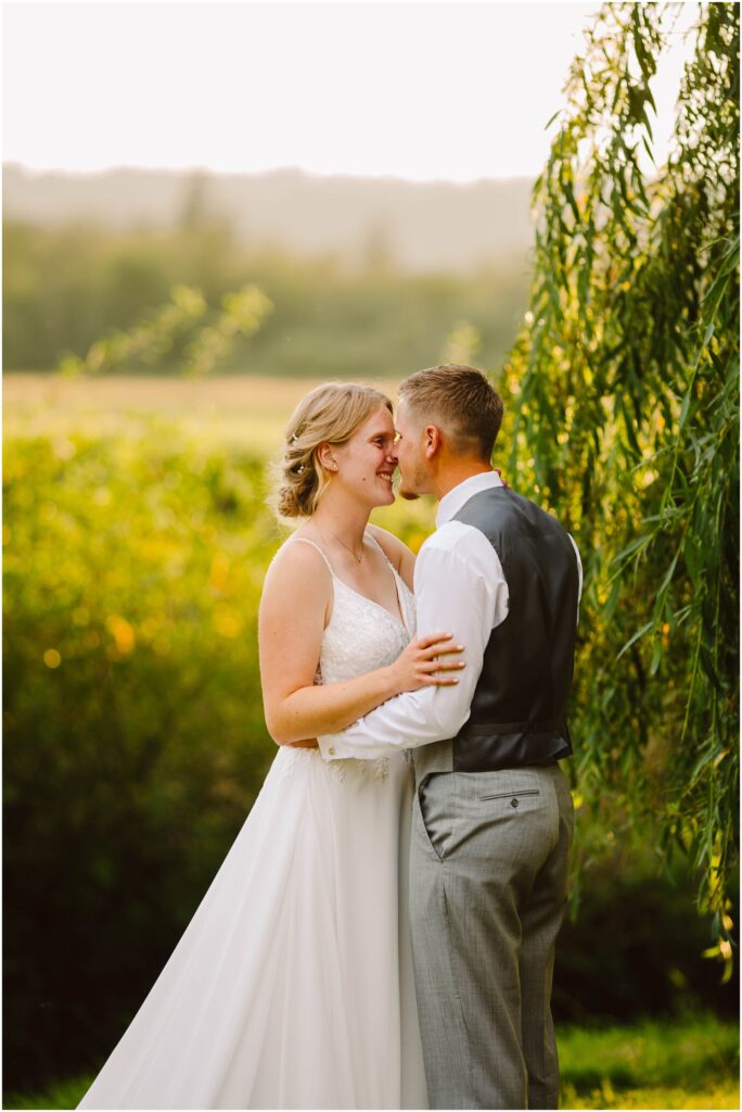Bride and groom standing close under a willow tree smiling at each other in golden evening light Snohomish wedding photographer gsquared weddings a couple stands outdoors in a lush garden setting during sunset The woman wears a white wedding dress and the man is in a shirt vest and pants They embrace and smile at each other surrounded by greenery and soft warm light Serving seattle snohomish and washington state