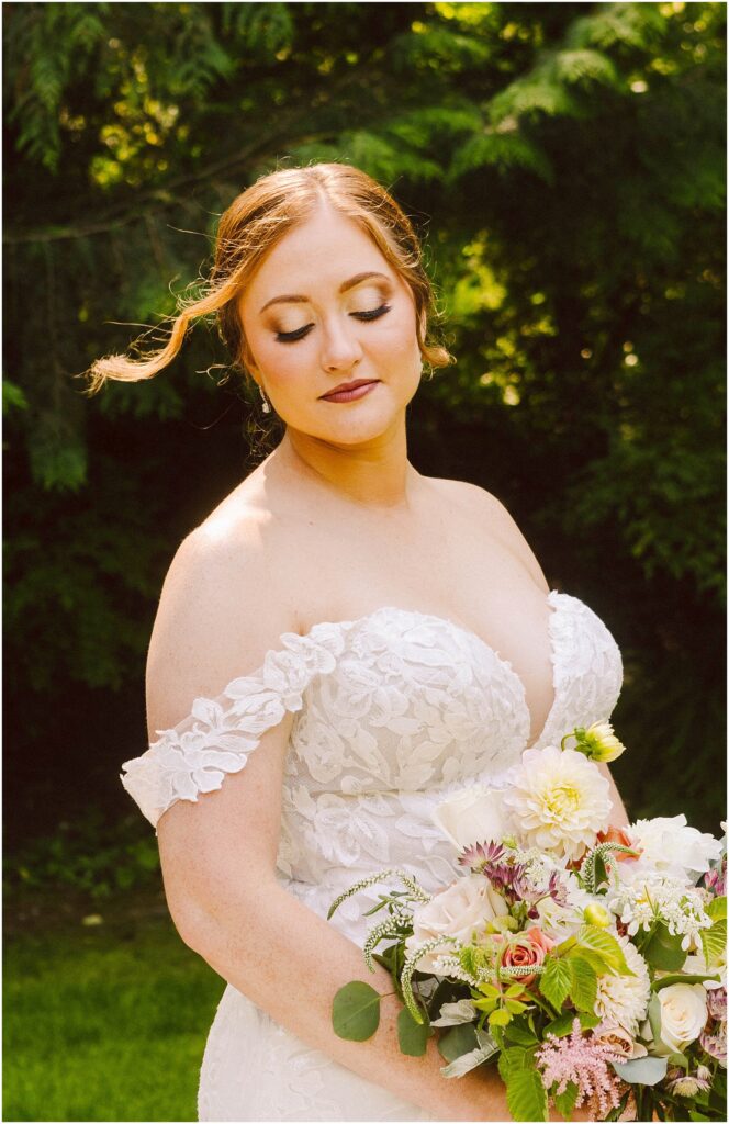 Bride in an off-shoulder lace gown holding a bouquet of roses and dahlias eyes closed in soft sunlight Snohomish wedding photographer gsquared weddings bride in an off shoulder lace wedding dress holds a bouquet of mixed flowers She stands outdoors against a backdrop of lush greenery with her eyes closed and a serene expression as a wisp of hair gently falls across her face Serving seattle snohomish and washington state