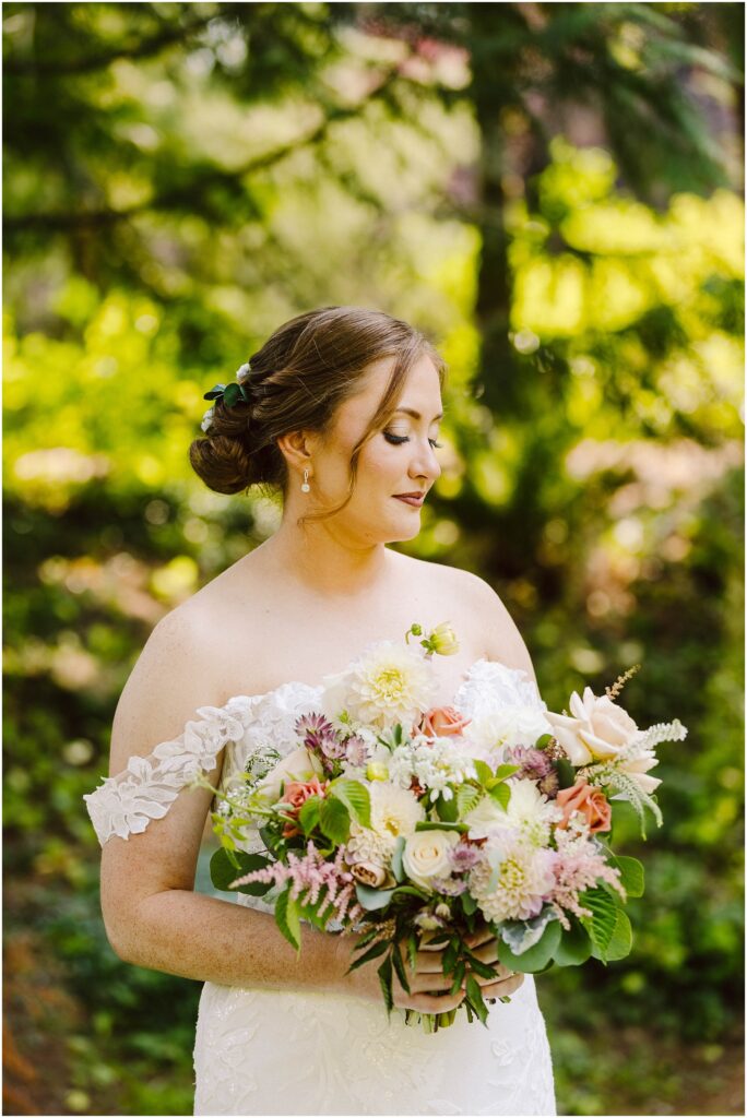Snohomish wedding photographer gsquared weddings a bride in a lace off-shoulder wedding dress stands outdoors holding a colorful bouquet Her hair is styled in an updo adorned with greenery The background is a lush sunlit garden with green foliage Serving seattle snohomish and washington state Snohomish wedding photographer gsquared weddings a bride in a lace off shoulder wedding dress stands outdoors holding a colorful bouquet Her hair is styled in an updo adorned with greenery The background is a lush sunlit garden with green foliage Serving seattle snohomish and washington state