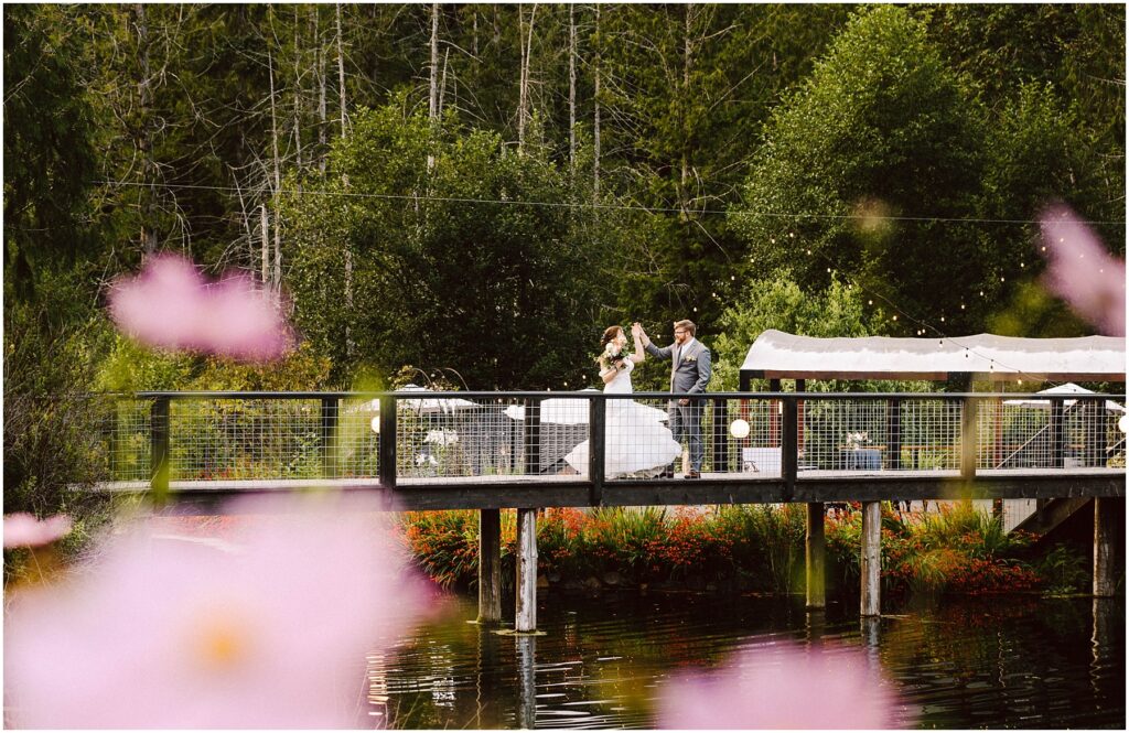 Snohomish wedding photographer gsquared weddings a bride and groom walk across a wooden bridge over a pond surrounded by lush greenery and colorful flowers The background features tall trees and soft pink flowers are in the foreground partially framing the scene Serving seattle snohomish and washington state Snohomish wedding photographer gsquared weddings a bride and groom walk across a wooden bridge over a pond surrounded by lush greenery and colorful flowers The background features tall trees and soft pink flowers are in the foreground partially framing the scene Serving seattle snohomish and washington state
