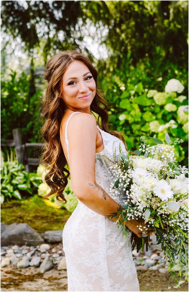 Snohomish wedding photographer gsquared weddings a bride in a white lace wedding dress stands outdoors holding a bouquet of white flowers and greenery She has long wavy brown hair and is smiling slightly The background features lush greenery and a wooden fence Serving seattle snohomish and washington state Snohomish wedding photographer gsquared weddings a bride in a white lace wedding dress stands outdoors holding a bouquet of white flowers and greenery She has long wavy brown hair and is smiling slightly The background features lush greenery and a wooden fence Serving seattle snohomish and washington state