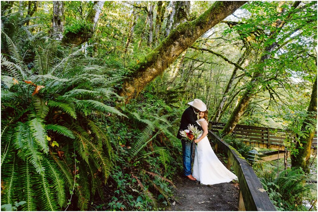 Snohomish wedding photographer gsquared weddings a bride in a white dress and a groom in a cowboy hat embrace on a forest path surrounded by lush green ferns and tall trees A wooden bridge is visible in the background under dappled sunlight Serving seattle snohomish and washington state Snohomish wedding photographer gsquared weddings a bride in a white dress and a groom in a cowboy hat embrace on a forest path surrounded by lush green ferns and tall trees A wooden bridge is visible in the background under dappled sunlight Serving seattle snohomish and washington state