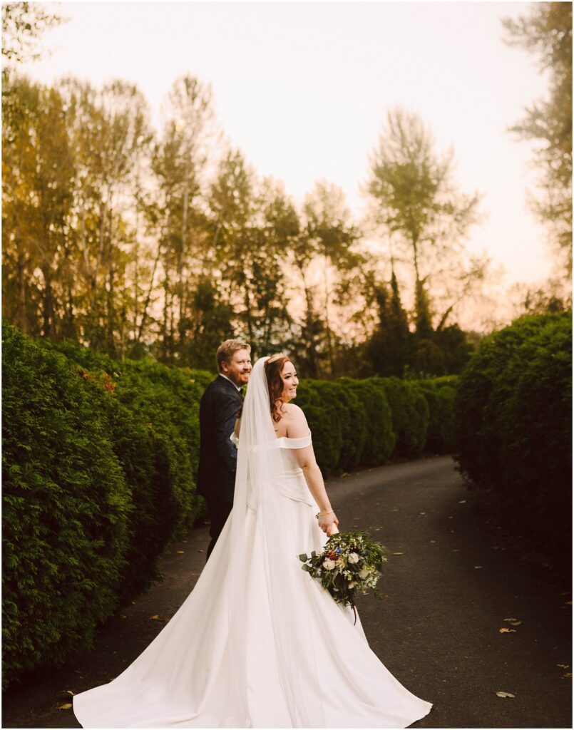 Snohomish wedding photographer gsquared weddings bride in an off-shoulder gown and veil holds a bouquet while walking hand-in-hand with a groom in a dark suit They stroll on a tree-lined path at sunset looking back and smiling Serving seattle snohomish and washington state Snohomish wedding photographer gsquared weddings bride in an off shoulder gown and veil holds a bouquet while walking hand in hand with a groom in a dark suit They stroll on a tree lined path at sunset looking back and smiling Serving seattle snohomish and washington state
