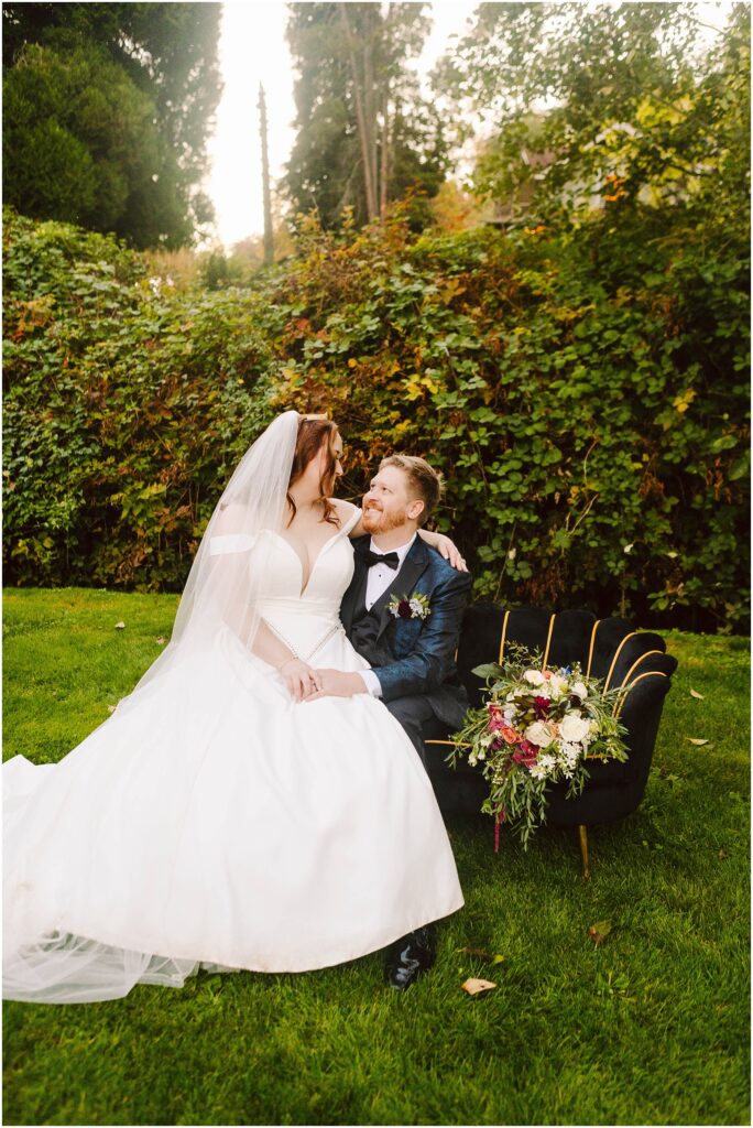 Snohomish wedding photographer gsquared weddings a bride in a white dress and veil sits beside a groom in a black suit and bowtie on a chair outdoors They are smiling at each other A vibrant bouquet is placed on the chair There is greenery in the background Serving seattle snohomish and washington state Snohomish wedding photographer gsquared weddings a bride in a white dress and veil sits beside a groom in a black suit and bowtie on a chair outdoors They are smiling at each other A vibrant bouquet is placed on the chair There is greenery in the background Serving seattle snohomish and washington state