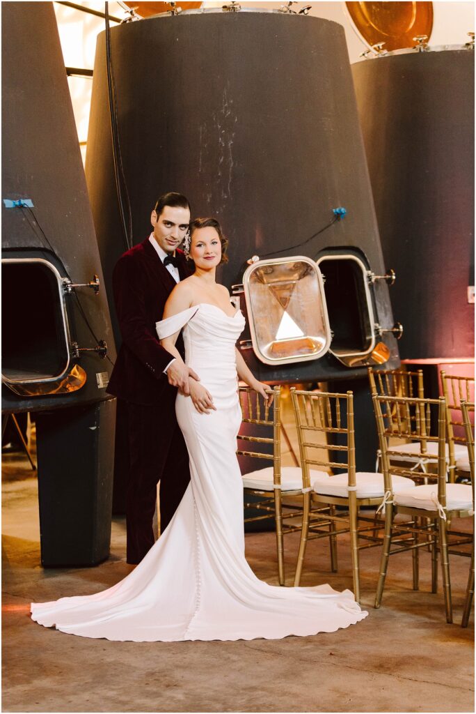 Snohomish Wedding Photographer GSquared Weddings A couple stands in front of large industrial tanks. The woman wears an off-shoulder white gown, and the man wears a burgundy suit. They are surrounded by rows of chairs, suggesting a wedding or formal event setting. Serving Seattle, Snohomish and Washington State