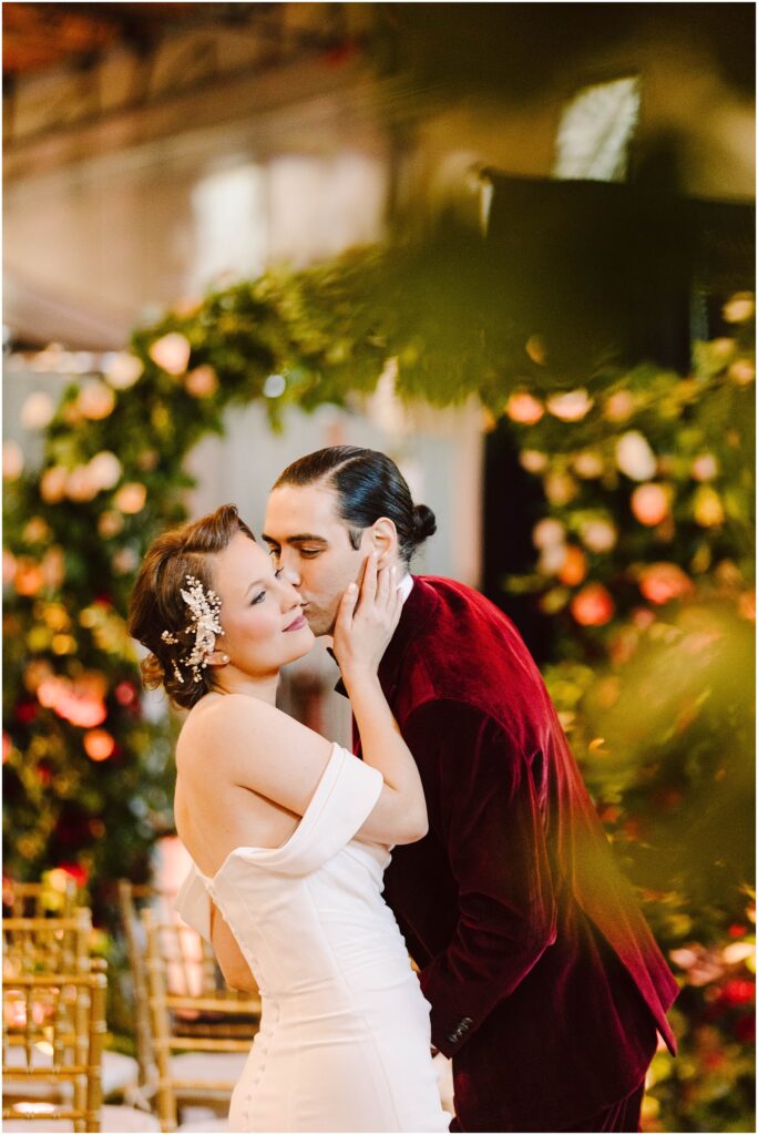 Snohomish Wedding Photographer GSquared Weddings A couple shares a tender moment at a wedding ceremony. The woman wears a white dress with off-the-shoulder sleeves and a decorative hairpiece, while the man wears a red velvet suit. They are surrounded by lush greenery and floral arrangements. Serving Seattle, Snohomish and Washington State