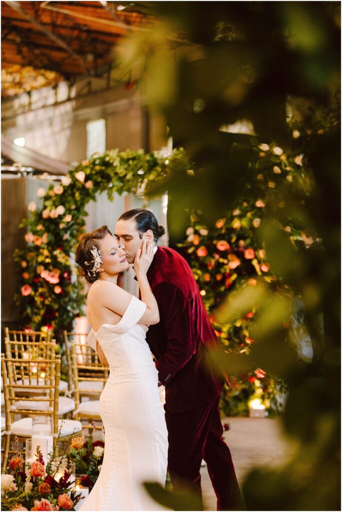 Snohomish Wedding Photographer GSquared Weddings A couple shares an intimate moment during their wedding. The bride is in a white gown with floral hair accessories, while the groom wears a burgundy suit. Theyre surrounded by floral decorations and gold chairs. Serving Seattle, Snohomish and Washington State