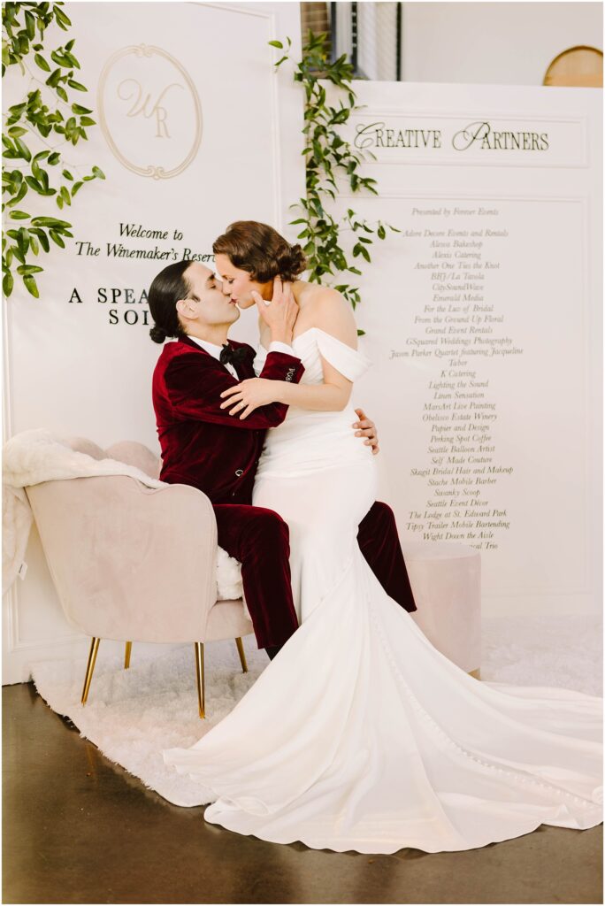 Snohomish Wedding Photographer GSquared Weddings A couple in wedding attire shares a kiss. The person in a white gown is seated on a plush chair, while the person in a burgundy suit kneels beside them. Behind them is a decorated panel with writing and green foliage. Serving Seattle, Snohomish and Washington State