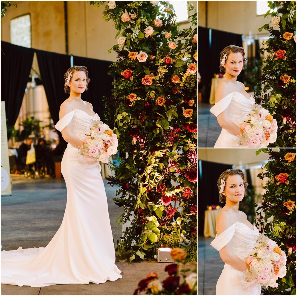 Snohomish Wedding Photographer GSquared Weddings A bride in an off-shoulder white dress holds a bouquet of pastel flowers. She stands beside a tall floral arrangement with roses and greenery. The setting is indoors with dark drapery and warm lighting. Serving Seattle, Snohomish and Washington State