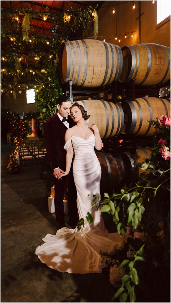 Snohomish Wedding Photographer GSquared Weddings A couple stands in a romantic winery setting. The woman is in a white gown, and the man is in a dark suit. They hold hands near a stack of wooden barrels, surrounded by warm lighting and lush greenery. Serving Seattle, Snohomish and Washington State