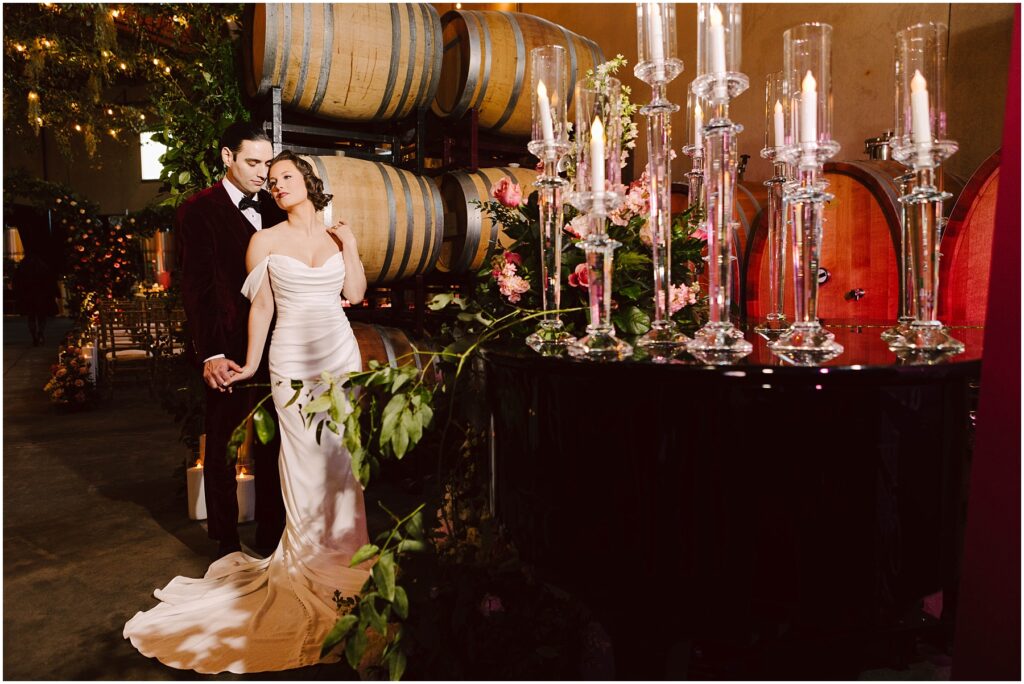 Snohomish Wedding Photographer GSquared Weddings A couple in formal attire stands beside a black grand piano adorned with flowers and lit candles. Behind them are wine barrels and greenery, creating a romantic ambiance. The setting appears to be a winery or cellar. Serving Seattle, Snohomish and Washington State