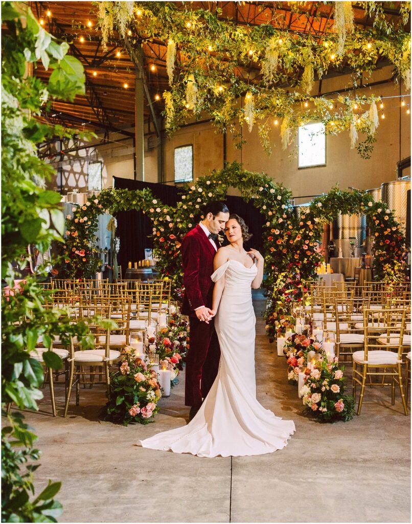 Snohomish Wedding Photographer GSquared Weddings A couple stands close together in a wedding venue, surrounded by gold chairs and vibrant floral arrangements. The bride wears an elegant white gown, and the groom is in a red velvet suit. Greenery and string lights are suspended above them. Serving Seattle, Snohomish and Washington State