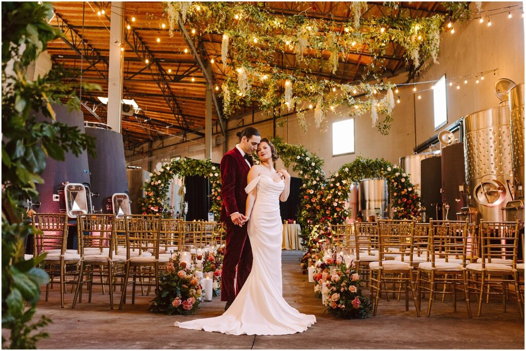 Snohomish Wedding Photographer GSquared Weddings A bride and groom stand in an elegantly decorated industrial venue. The groom wears a burgundy suit, and the bride wears a white dress. There are floral arrangements and chairs arranged for an event. String lights and greenery hang overhead. Serving Seattle, Snohomish and Washington State