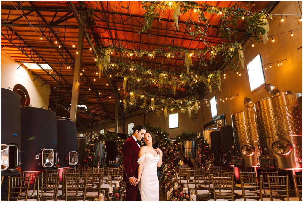 Snohomish Wedding Photographer GSquared Weddings A bride in a white dress and a groom in a burgundy suit stand close in a warmly lit industrial venue decorated with greenery and string lights. Rows of chairs and large metal tanks are visible in the background. Serving Seattle, Snohomish and Washington State