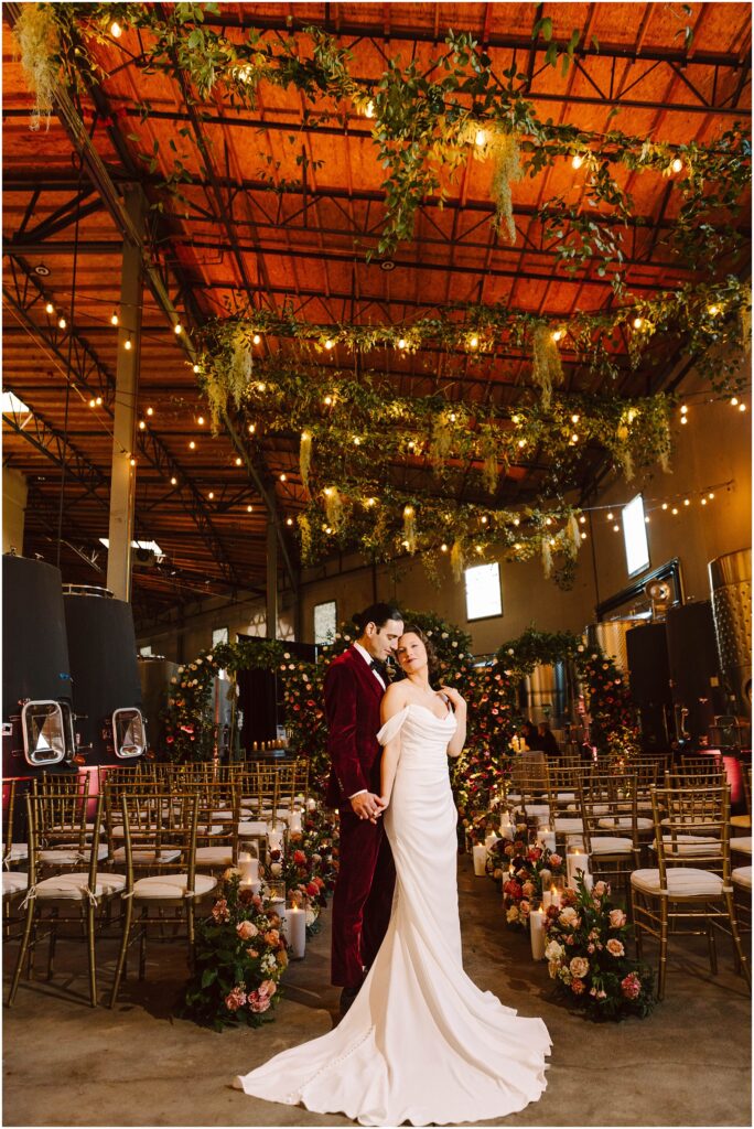 Snohomish Wedding Photographer GSquared Weddings A couple poses in a decorated indoor wedding venue. The bride wears a white gown, and the groom dons a red suit. The space is adorned with hanging greenery, string lights, and abundant floral arrangements, with rows of chairs for guests. Serving Seattle, Snohomish and Washington State