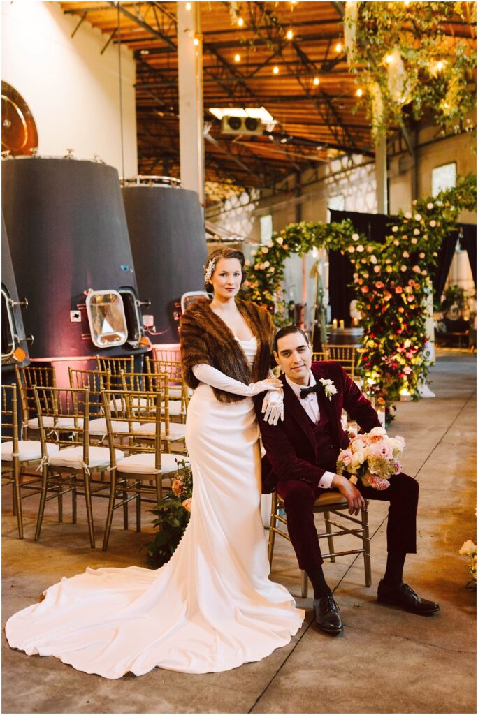 Snohomish Wedding Photographer GSquared Weddings A couple in vintage attire poses in a beautifully decorated venue. The woman in a white gown and fur stole stands beside the seated man in a burgundy suit. Floral arrangements and rows of chairs are visible in the background. Serving Seattle, Snohomish and Washington State