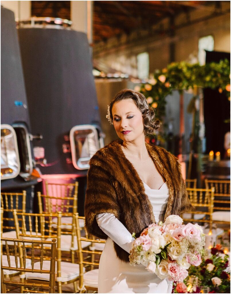 Snohomish Wedding Photographer GSquared Weddings A woman in a fur shawl holds a bouquet of pink and white flowers. She stands in a venue with rows of gold chairs, and large wine tanks are visible in the background. The setting suggests a rustic and elegant event atmosphere. Serving Seattle, Snohomish and Washington State
