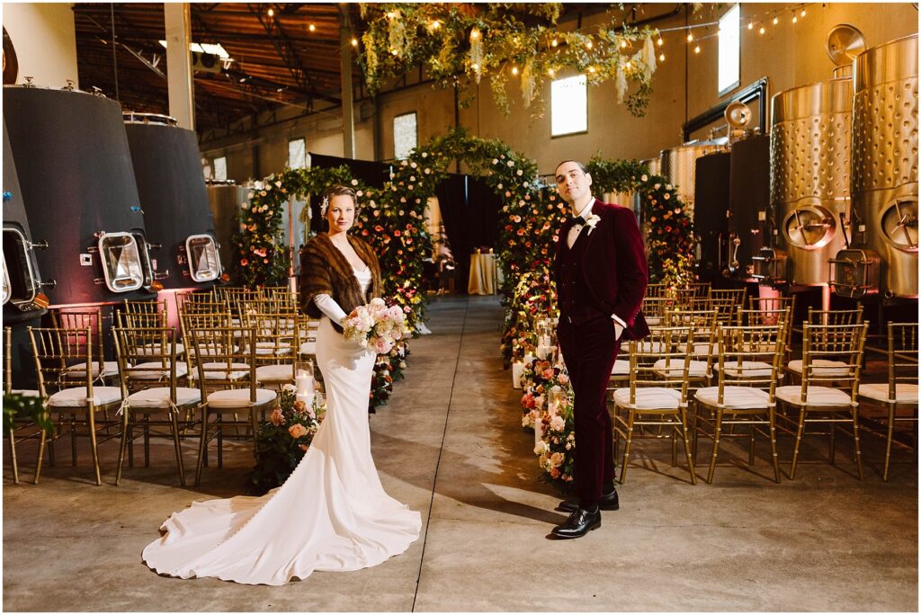 Snohomish Wedding Photographer GSquared Weddings A bride and groom stand in the center of an aisle adorned with flowers in an industrial-style venue. She wears a white gown and fur wrap, holding a bouquet. He wears a burgundy suit. Rows of gold chairs and string lights enhance the setting. Serving Seattle, Snohomish and Washington State
