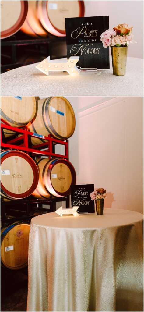 Snohomish Wedding Photographer GSquared Weddings A small round table with a gold tablecloth holds a lit arrow sign, a black sign with cursive text, and a vase with pink and peach flowers. Behind the table are stacked wooden barrels. Serving Seattle, Snohomish and Washington State