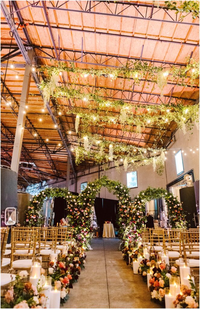 Snohomish Wedding Photographer GSquared Weddings A beautifully decorated indoor wedding aisle with two floral arches adorned with greenery and colorful flowers. Gold chairs line the aisle, and candles in glass holders are placed on the floor. Hanging greenery decorates the ceiling, enhancing the warm ambiance. Serving Seattle, Snohomish and Washington State
