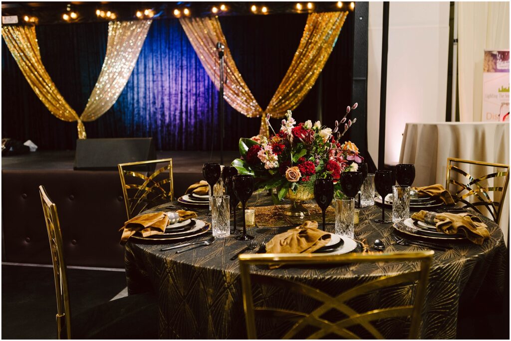 Snohomish Wedding Photographer GSquared Weddings Elegant event table setting with gold and black theme. Features gold chairs, a patterned tablecloth, black and gold dinnerware, and a centerpiece of vibrant flowers. In the background, a stage with gold drapes and string lights. Serving Seattle, Snohomish and Washington State