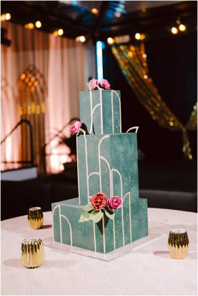 Snohomish Wedding Photographer GSquared Weddings A tall, green, three-tiered cake with white geometric patterns and pink roses on top. The cake sits on a table adorned with a textured white cloth and surrounded by small golden candle holders. The background features soft lighting and elegant drapes. Serving Seattle, Snohomish and Washington State