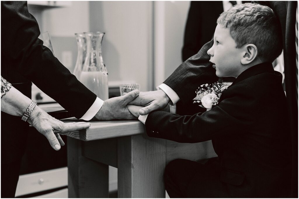 Snohomish Wedding Photographer GSquared Weddings A black and white photo of a young boy in a suit sitting at a table. Several adult hands are placed on top of each other next to him, symbolizing unity or support. A glass pitcher is visible in the background. Serving Seattle, Snohomish and Washington State