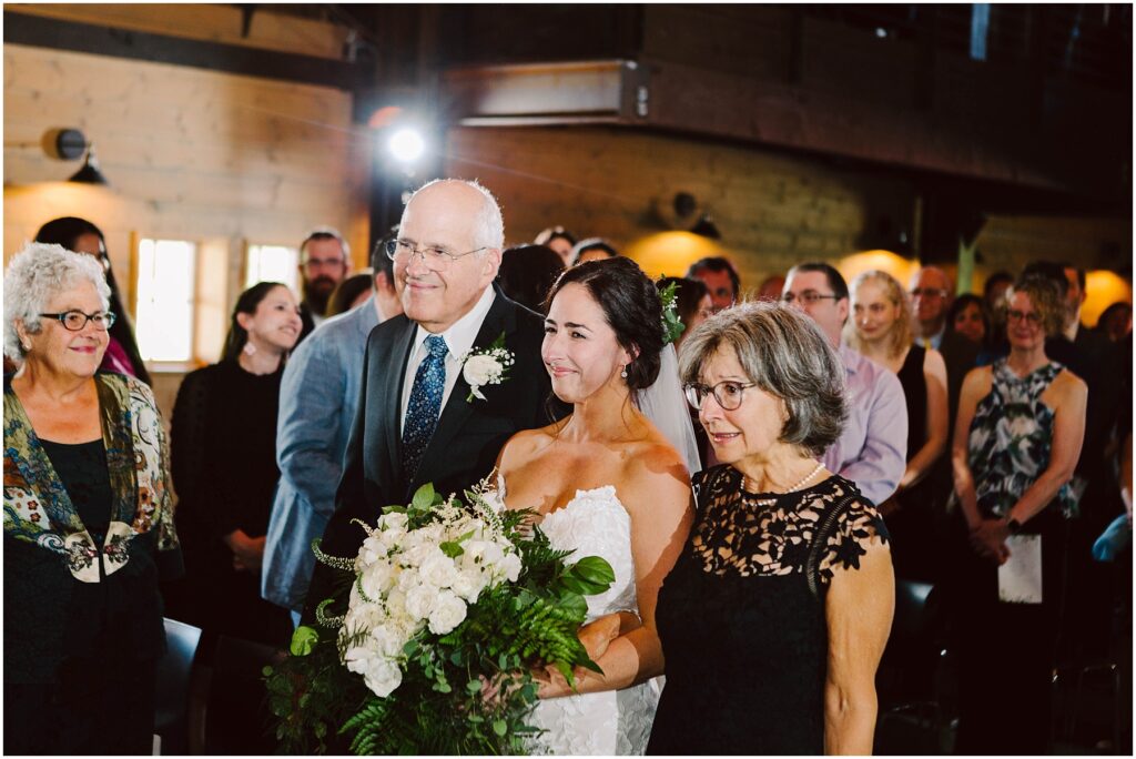 Snohomish Wedding Photographer GSquared Weddings A bride in a white dress holding a bouquet of white flowers stands between two older adults, smiling as they walk down an aisle. Guests are seated on both sides, watching the procession in a warmly lit venue. Serving Seattle, Snohomish and Washington State
