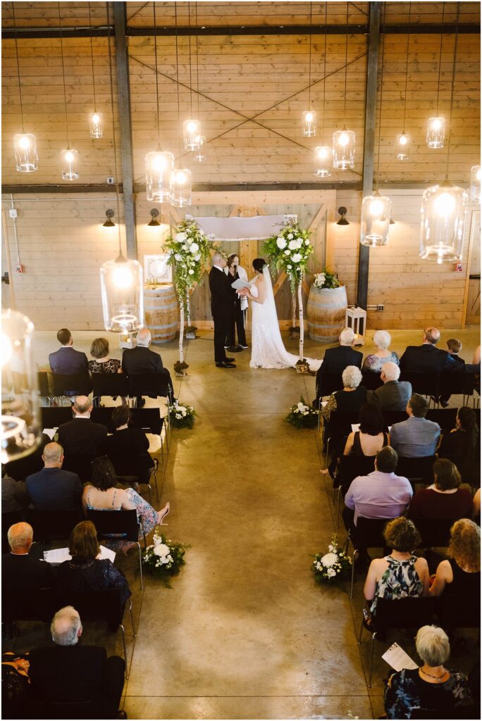 Snohomish Wedding Photographer GSquared Weddings A wedding ceremony in a rustic venue with wooden walls. The couple stands at the altar adorned with flowers and barrels, surrounded by seated guests. Overhead, warm hanging lights illuminate the scene. Serving Seattle, Snohomish and Washington State