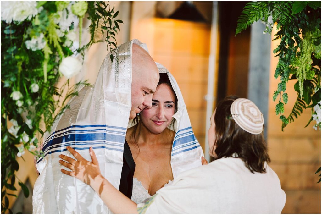 Snohomish Wedding Photographer GSquared Weddings A couple, covered with a white and blue-striped cloth, stands closely together under a canopy. They are joined by a person facing them, wearing a kippah. The setting is adorned with greenery and white flowers, and the atmosphere is serene and intimate. Serving Seattle, Snohomish and Washington State